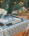 A styled outdoor mahjong table setup with a blue and white floral patterned leatherette mahjong mat, a scalloped tortoiseshell rack set with  mahjong tiles bamboo chairs in a garden setting with warm string lights.