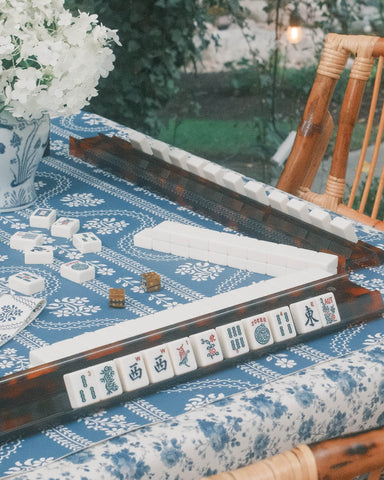 A mahjong set displayed on a blue and white patterned tablecloth in an outdoor garden setting with wooden chairs and warm string lights in the background.