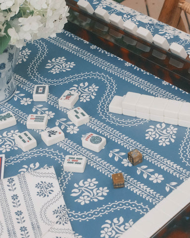 A mahjong game setup on a blue and white serpentine patterned leatherette mahjong mat, coordinating  tablecloth, with white tiles arranged on scalloped tortoiseshell racks.  A red National Mahjong League card is visible in the lower left, along with a vase of white flowers.