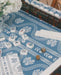 A mahjong game setup on a blue and white serpentine patterned leatherette mahjong mat, coordinating  tablecloth, with white tiles arranged on scalloped tortoiseshell racks.  A red National Mahjong League card is visible in the lower left, along with a vase of white flowers.