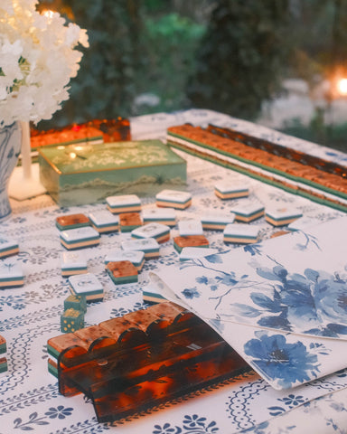 A styled table setting featuring a blue and white floral leatherette mahjong mat and tablecloth, with scalloped tortoiseshell mahjong racks.
