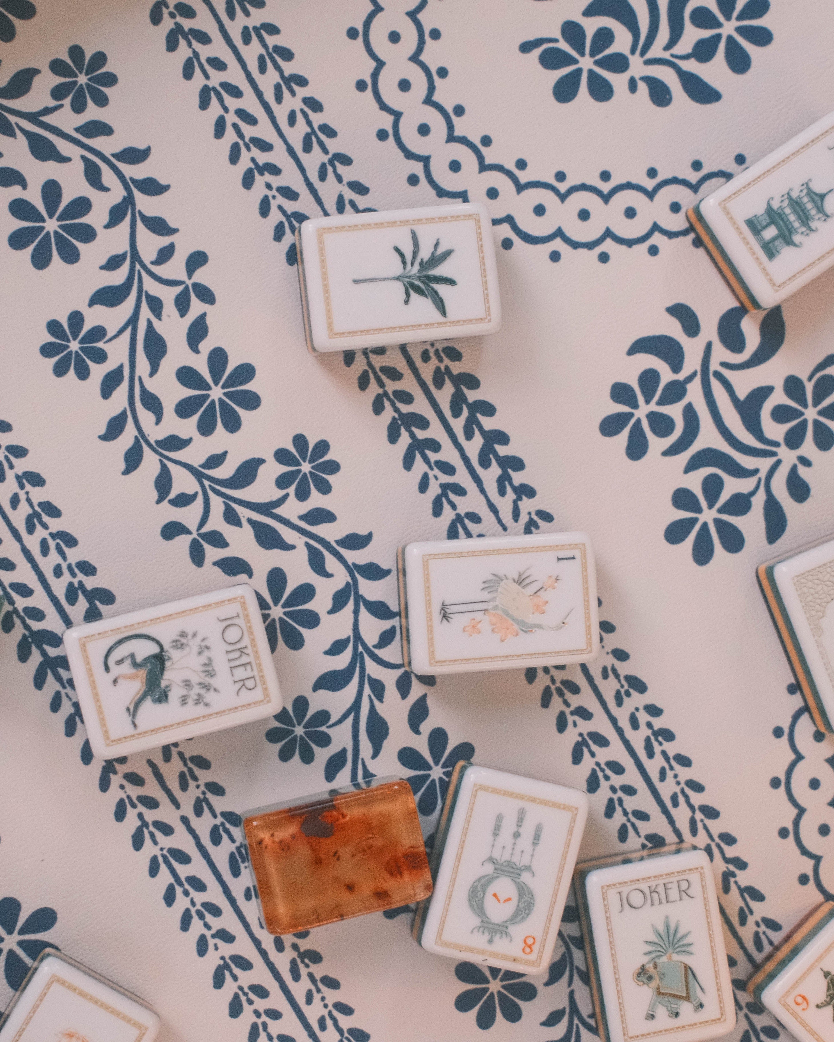 A styled flat-lay of a mahjong game, showing several mahjong tiles with botanical illustrations and two green dice on a cream-colored serpentine pattern leatherette mat with a blue floral serpentine pattern.