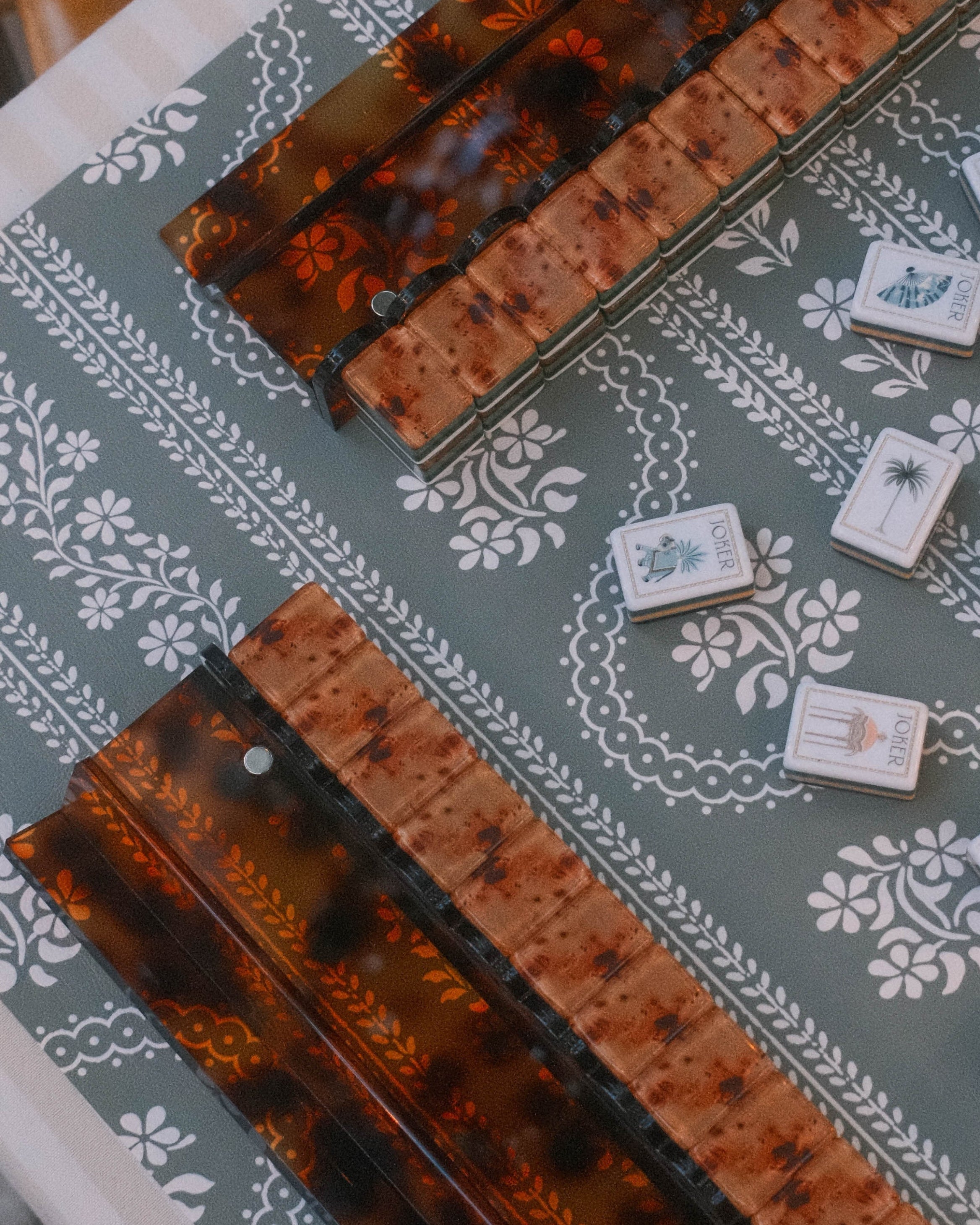 A close-up overhead view of a mahjong table setup with two 20' tortoiseshell scalloped mahjong racks on a green floral serpentine leatherette mahjong mat with mahjong tiles.