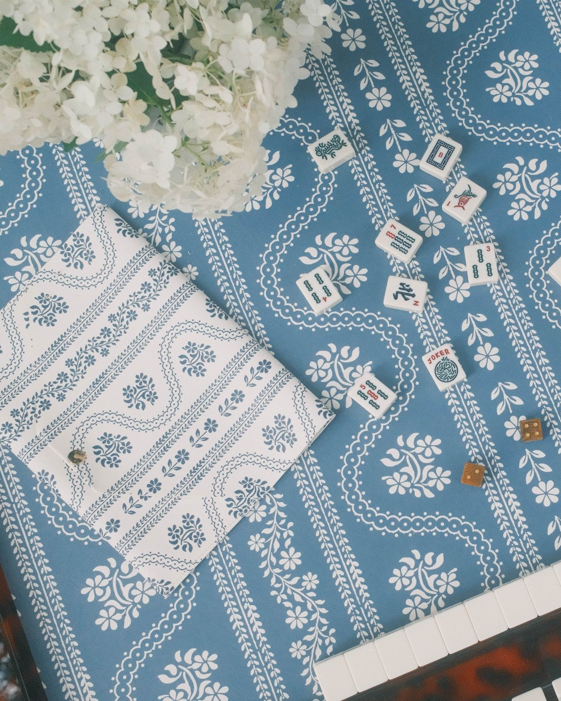 A blue and white patterned mahjong mat with a bohemian-style floral and paisley design, styled on a wooden table with mahjong tiles, a folded matching napkin, and a rattan chair partially visible.