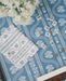 A blue and white patterned mahjong mat with a bohemian-style floral and paisley design, styled on a wooden table with mahjong tiles, a folded matching napkin, and a rattan chair partially visible.