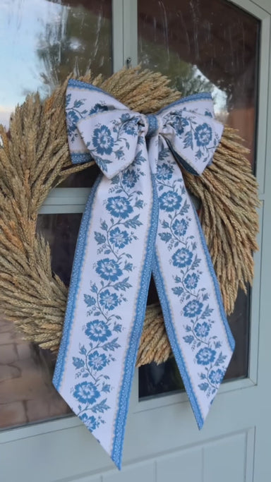 Natural wheat wreath adorned with an oversized blue and white floral bow, styled on a glass door for a warm fall entrance.
